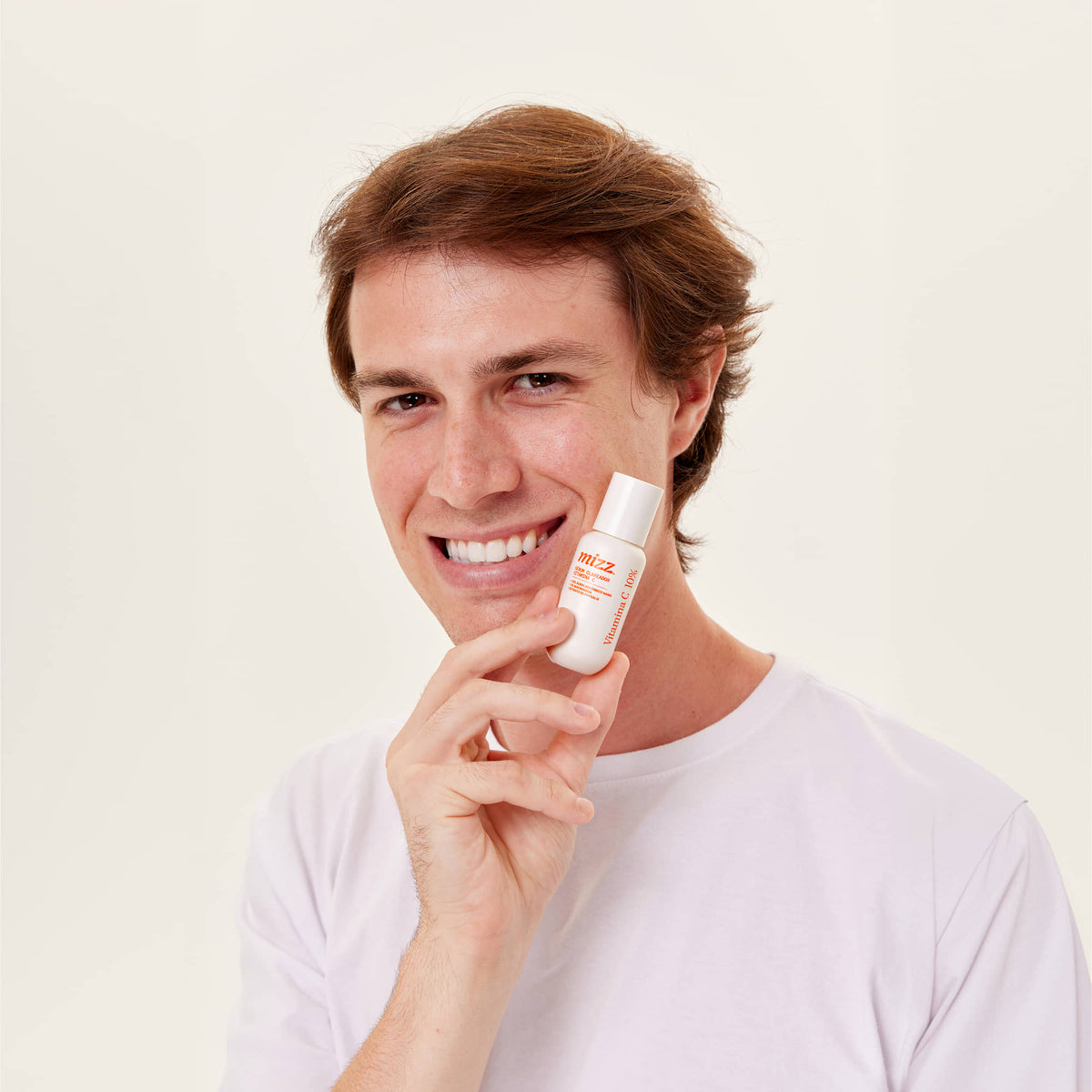 Jovem sorrindo de camiseta branca segurando Sérum Vitamina C 10% Mizz com detalhes laranja